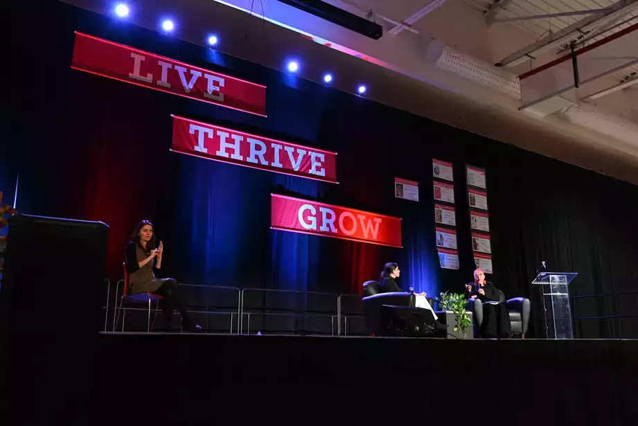 Mills Distinguished Lecture host, guest, and ASL interpreter seen on the stage with "Grow, Thrive, Live" seen in the background